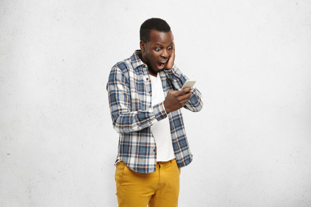 African American black man shocked, holding smart phone in one hand touching his head Free Photo