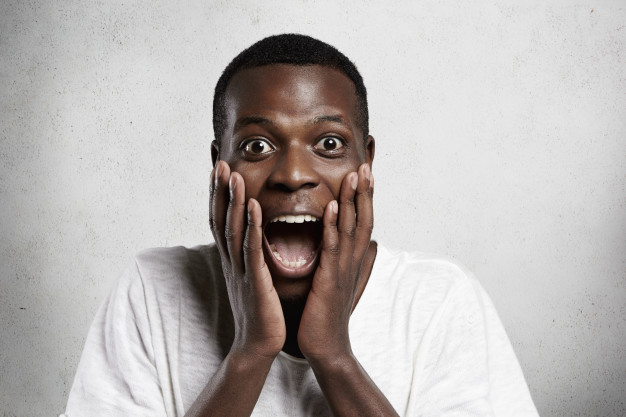 African employee or customer with shocked and surprised face, looking and screaming with big eyes and mouth wide open, holding hands on his cheeks. Free Photo