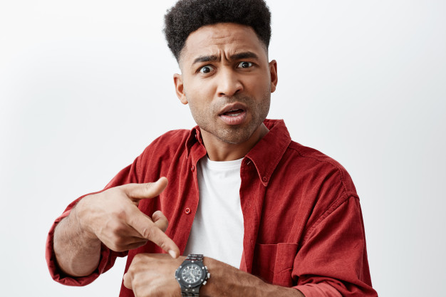 Close up portrait of young handsome black-skinned man with afro hairstyle in casual white t-shirt under red shirt pointing at hand watch with unsatisfied expression, after his friend being late. Free Photo