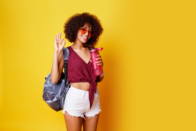 Graceful sport black female standing on yellow and holding pink bottle of water wearing stylish summer clothes and back pack. Free Photo