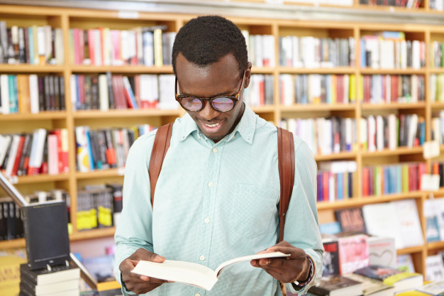 Handsome young afro american hipster in shades holding open book in his hands, reading his favorite poem, searching for inspiration in public library or bookstore. people, lifestyle and leisure Free Photo