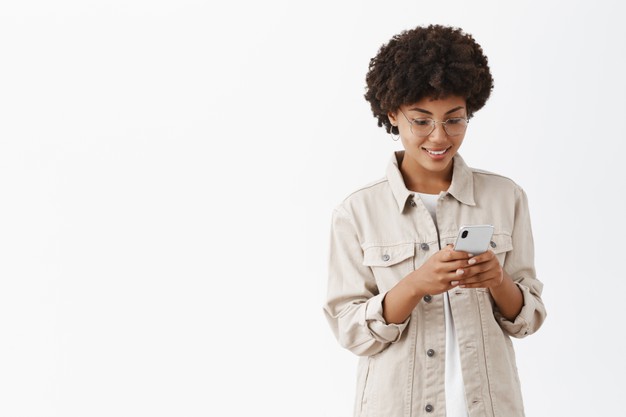 African American black woman happy young woman in glasses with afro hairstyle typing message in smartphone gazing with joyful smile at smartphone screen reading interesting article in web Free Photo