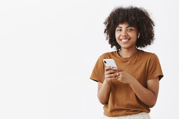 Modern stylish and pleased african american woman using wireless earphones while talking with clients holding smartphone making notes or picking music in device Free Photo