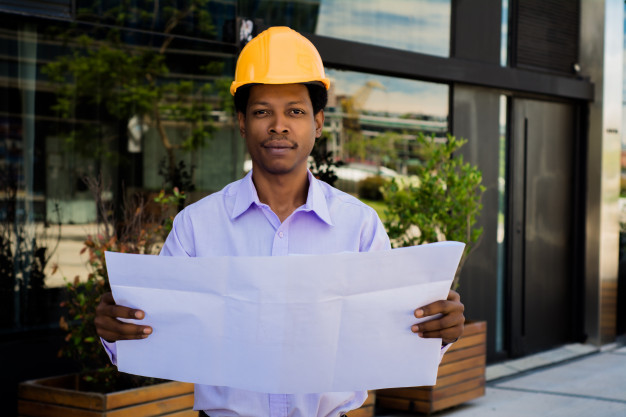 Portrait of professional architect in helmet looking at blue prints outside modern building. engineer and architect concept. Free Photo