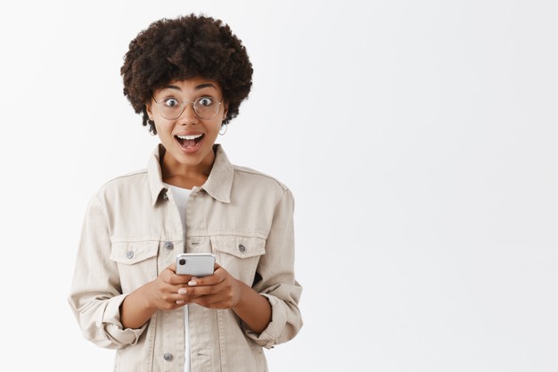 Stunned and amazed good-looking African American black boyish girl in beige shirt and glasses holding smartphone gasping and gazing with admiration and excitement Free Photo