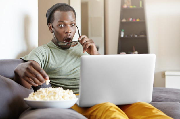 Surprised african male sitting on couch at home, eating popcorn and watching exciting tv show online on laptop computer or shocked with cliffhanger ending of detective series, keeping his mouth open Free Photo