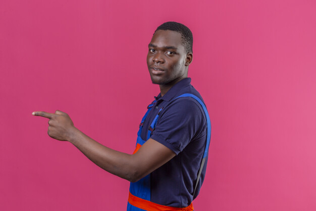 Young african american builder man wearing construction uniform standing sideways pointing to something on isolated pink Free Photo