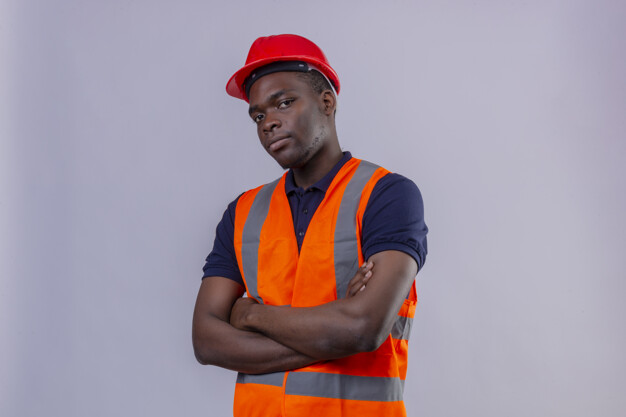 young african american builder man wearing construction vest and safety helmet standing with arms crossed looking suspicious over white background