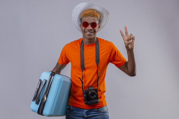 Young african american handsome traveler boy in summer hat wearing orange t-shirt holding travel suitcase looking at camera smiling friendly showing number two or victory sign over white backgrou Free Photo