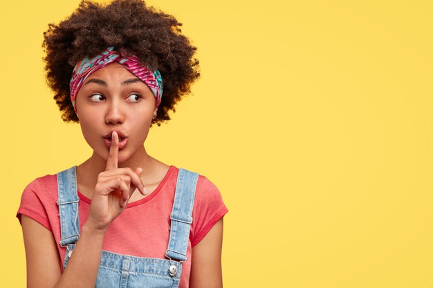 African american female with secret expression, makes silence sign, keeps fore finger on lips, looks aside, poses over yellow wall Free Photo