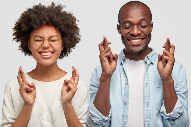 Cheerful african american couple cross fingers, have broad smiles, hope for good luck before exam, isolated over white wall. people, ethnicity and body language concept Free Photo