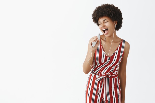 Indoor shot of creative and happy carefree african american woman with afro hairstyle holding smartphone like microphone singing along music while listening songs in earphones joyfully Free Photo