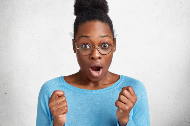 Scared dark skinned african female with curly hair, expresses amazement and shock, gestures actively and clenches fists from unexpectdeness Free Photo