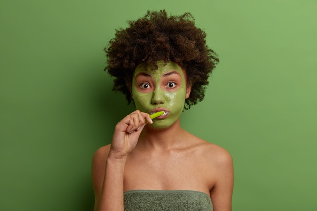 African American black woman or girl wears facial anti aging mask brushes teeth, wrapped in bath towel