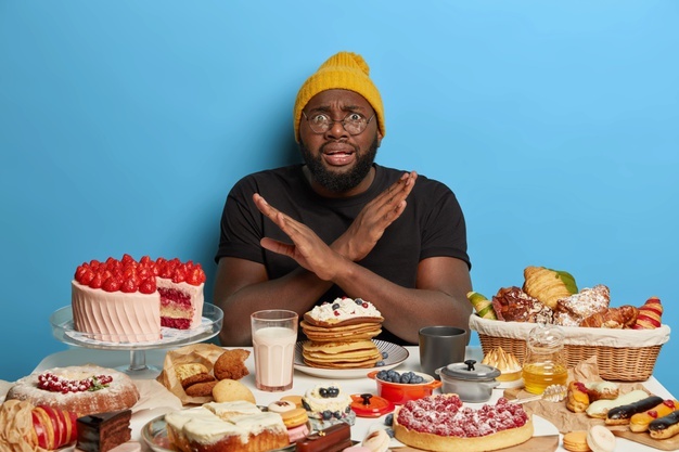 African american man crosses arms over chest, makes denial gesture, refuses eat sugary products, sits at table with bakery, keeps to diet Free Photo