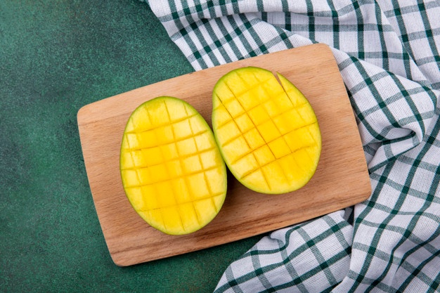 Top view of yellow delicious and exotic mango slices on a wooden kitchen board on checked tablecloth and green surface Free Photo