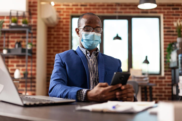 Entrepreneur looking at smartphone screen and wearing face mask at work in business office. company employee working on mobile phone with touch screen at desk during coronavirus pandemic. Free Photo