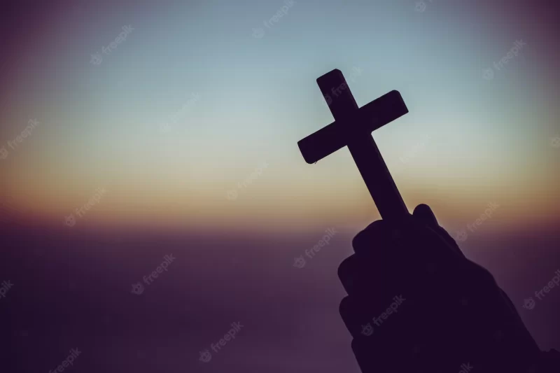 Silhouette of a man praying with a cross in hand at sunrise. Free Photo