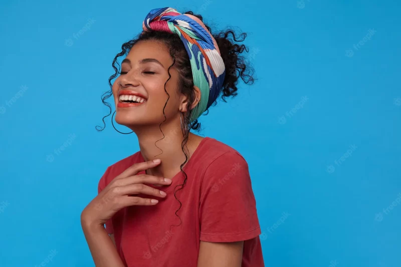 Indoor shot of young cheerful dark haired woman keeping raised hand on her chest and laughing happily with closed eyes, isolated over blue wall Free Photo