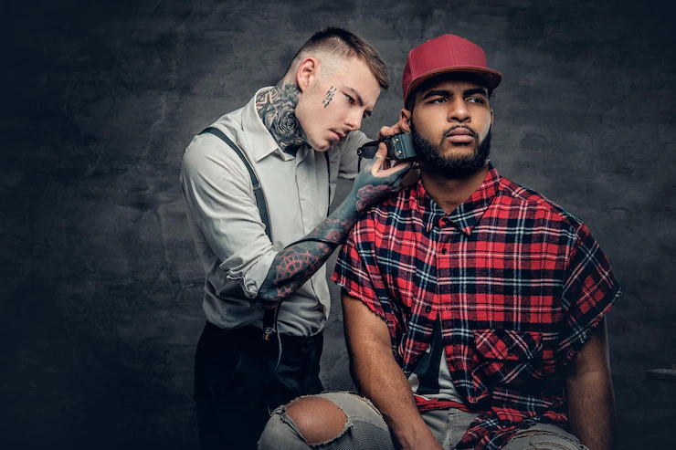 A tattooed caucasian male dressed in a white shirt cutting the beard of black hipster males in a red fleece shirt. Free Photo