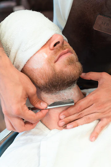 Vertical shot of a hairdresser shaving the beard of a young customer in a towel with a blade Free Photo