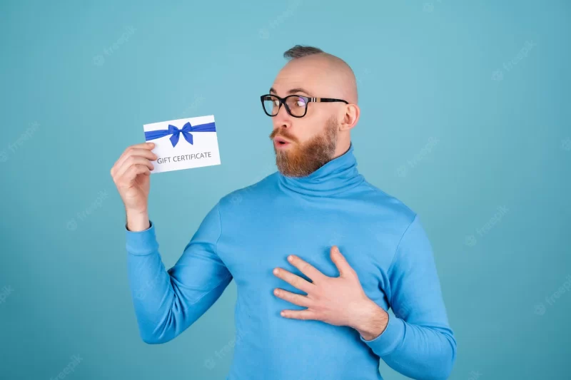 A young man with a red beard in a turtleneck and glasses on a blue background holds a gift certif