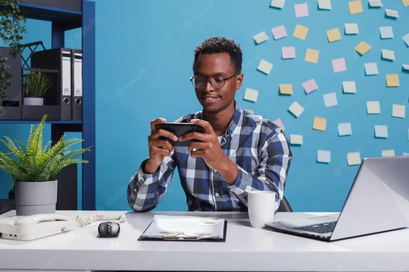 African american businessman playing games on mobile smartphone while in modern workspace office. marketing company financial advisor winning game on mobile device while at work. Free Photo