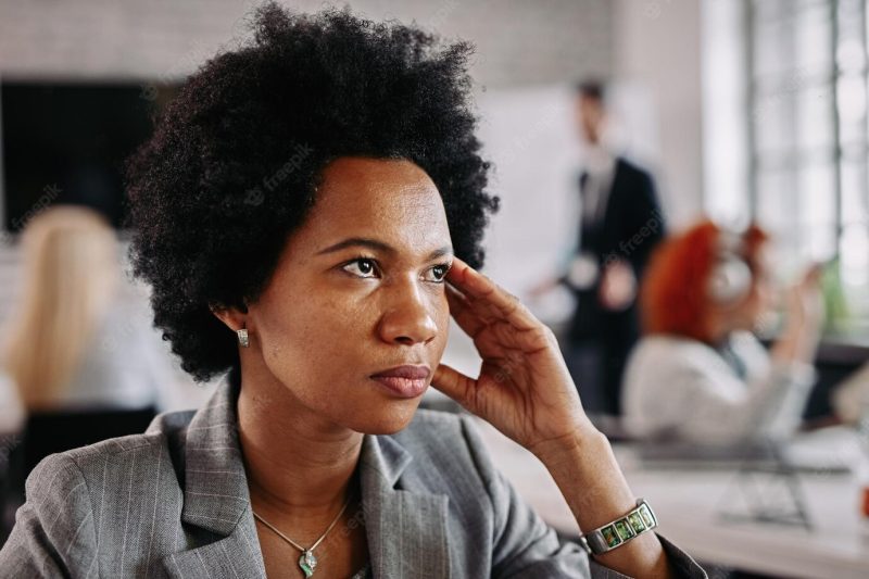 African American business woman looking thoughtful while being in the office there are people in the background Free Photo