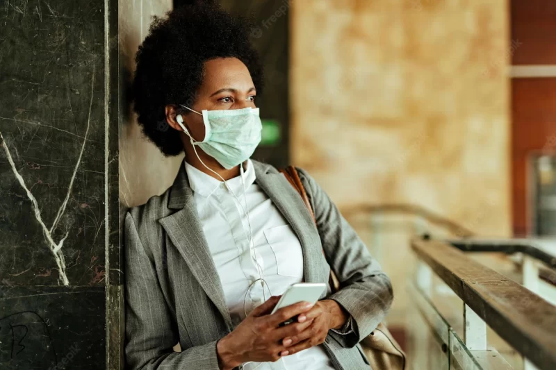 African american businesswoman wearing face mask for virus protection and using mobile phone while standing in a corridor Free Photo