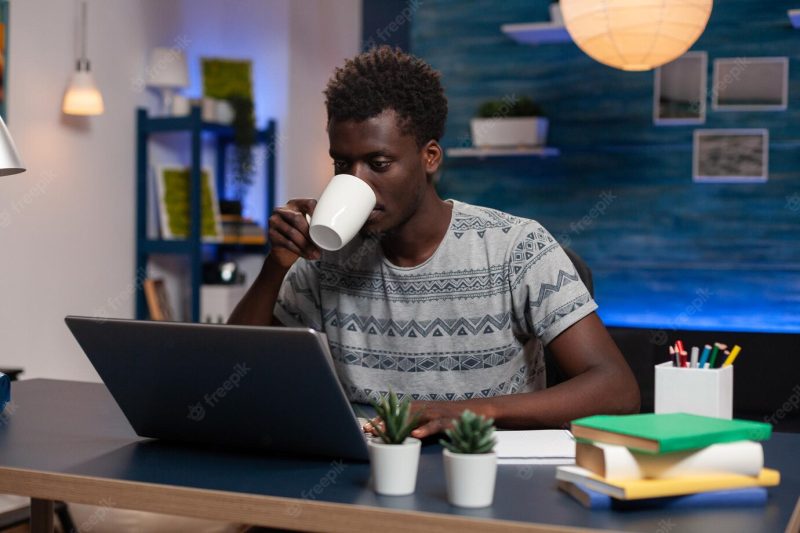 African american employee holding cup of coffee typing marketing ideas on computer working remote at online project in living room. freelancer sitting at desk analyzing financial graph Free Photo