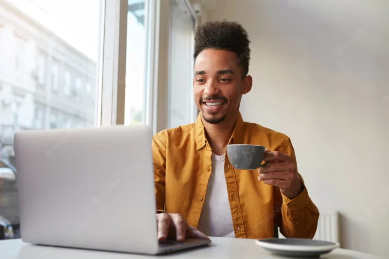 African American guy sitting at a table in a cafe and working on a laptop, wears in yellow shirt, drinks aromatic coffee, chatting with his girlfriend, enjoys the day. Free Photo