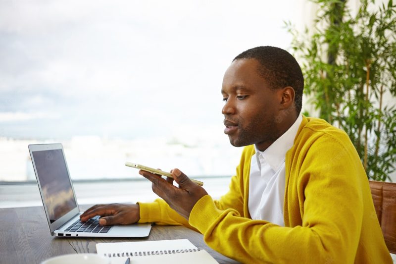 African American male freelancer sitting by large window at hotel lobby using wireless internet connection, working remotely on laptop and sending voice message via online app on mobile phone Free Photo