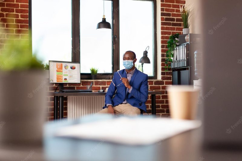 African american man wearing face mask and sitting in business office to work on strategy. company entrepreneur preparing to work on project planning during coronavirus pandemic. Free Photo