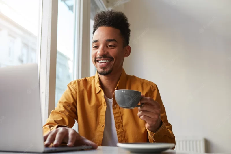 African American sitting at table in cafe and working laptop, wears in yellow shirt, drinks aromatic coffee, communicates with his sister which is far in another country , enjoys the work Free Photo