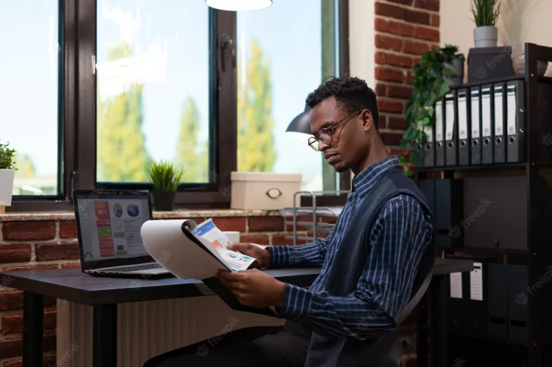 African american startup owner with glasses holding clipboard and paper with bussiness charts comparing data with sales results on laptop. entrepreneur analyzing marketing indicators on laptop. Free Photo