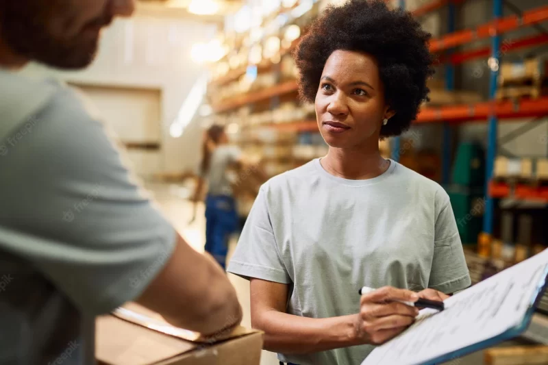 African american warehouse worker going through check list while communicating with her colleague in distribution warehouse Free Photo