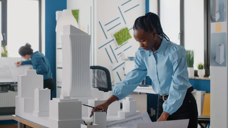 African american woman analyzing blueprints plan and building model to work on architecture. architect working with maquette and layout print to design urban construction for development. Free Photo