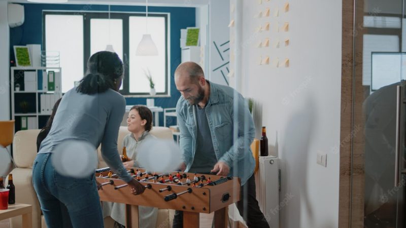 African american woman beating man at foosball game while enjoying after work drinks with workmates at office. colleagues playing soccer at football table to have fun and entertainment. Free Photo