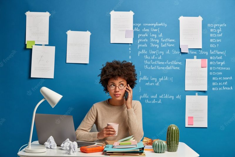 African american woman talks on mobile phone at workplace, chats with friend during coffee break, solves distance issues Free Photo