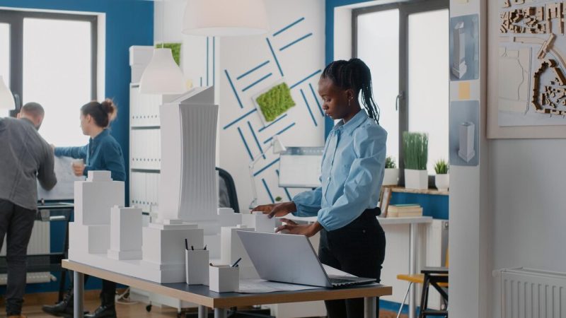 African american woman using laptop to design building model and blueprints plan. engineer working on computer to create architectural project with urban construction structure. Free Photo