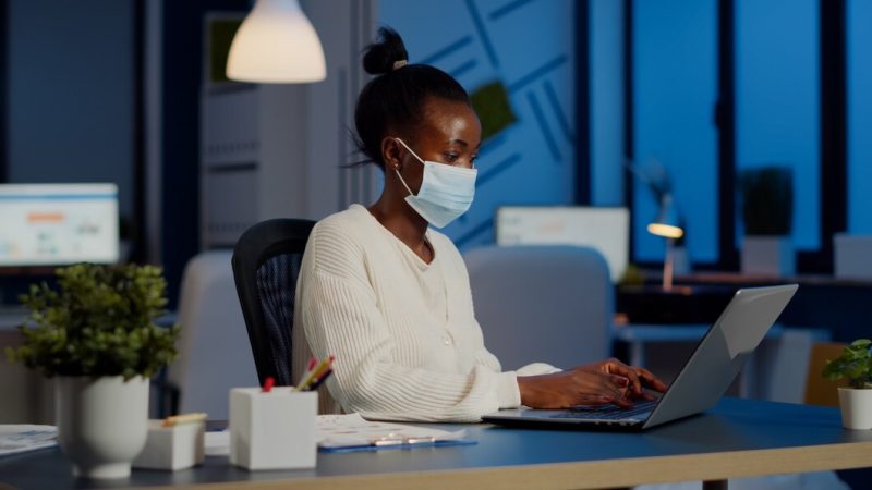 African employee with protection face mask working at laptop overtime in new normal business office to respect deadline of project, analysing documents sitting at desk overtime during global pandemic Free Photo