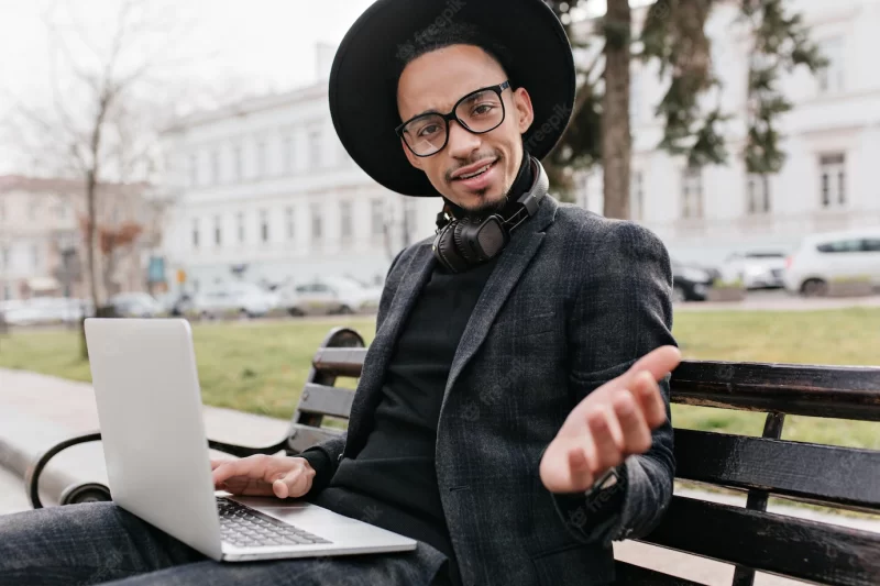 African freelancer expressing amazement while working with laptop in square. outdoor photo of surprised black man wears hat, sitting on bench and holding computer. Free Photo