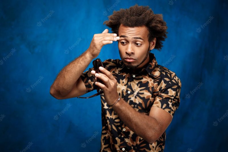 African man looking at reflection in sunglasses over blue surface Free Photo