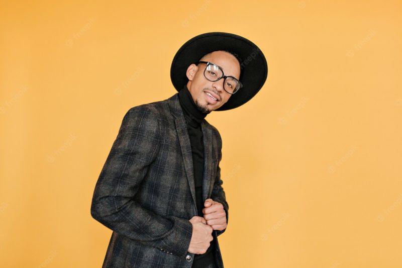 African man posing in gray checkered jacket. indoor portrait of funny mulatto male model standing beside yellow wall with interested face expression. Free Photo