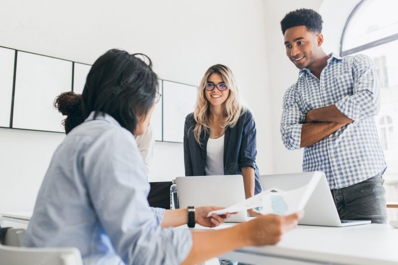 African office worker in checkered shirt standing with arms crossed and looking at asian manager. indoor portrait of freelance web-developers discussing something and using laptops. Free Photo