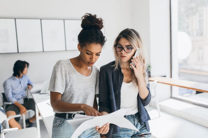 African woman in white t-shirt found mistake in the report. worried female office worker calling colleague to solve working problems while her co-workers doing their job. Free Photo