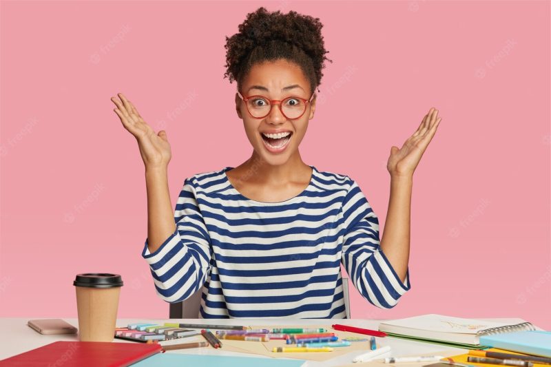 Amazed black female illustrator raises hands in eureka gesture, wears striped clothes Free Photo