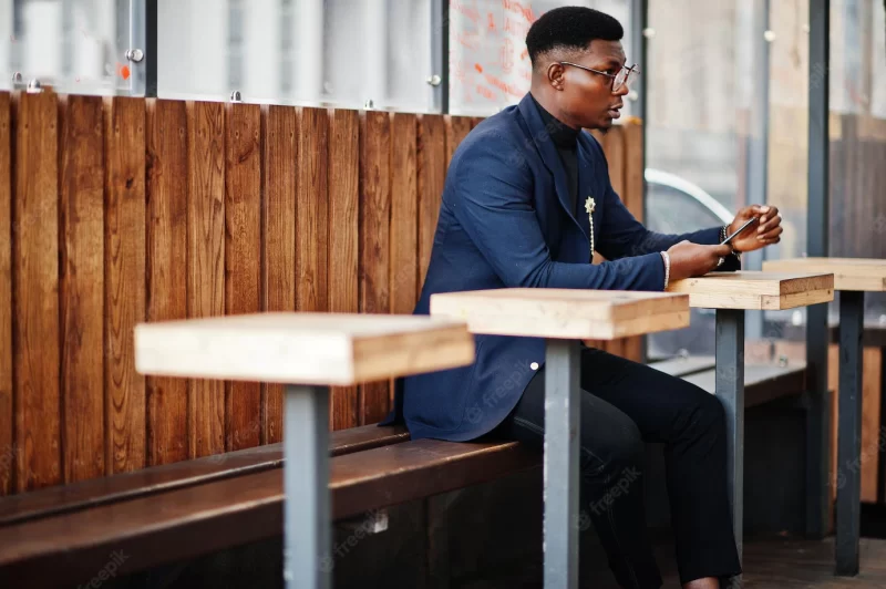 Amazingly looking african american man wear at blue blazer with brooch black turtleneck and glasses posed at street fashionable black guy sitting behind table with mobile phone Free Photo