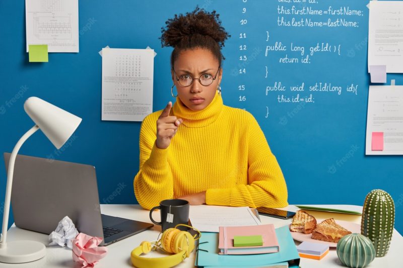 Annoyed afro american woman worker points at you and blames in doing something wrong, wears round glasses and yellow jumper, sits in co working space with mess on table. Free Photo