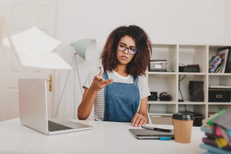 Annoyed curly girl in glasses and striped shirt scatter documents sitting at the table and looking away. unhappy young woman working with laptop and laptop doing her job in office. Free Photo